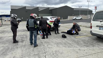 This photo shows a group of motorcyclists participating in a first aid training session in a car park. Several individuals wearing motorcycle gear and helmets are standing around, while others are actively involved in assisting a person lying on the ground.