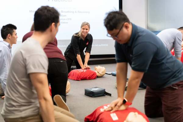 A trainer demonstrating CPR on manikins in the background, with students practicing in the foreground. 